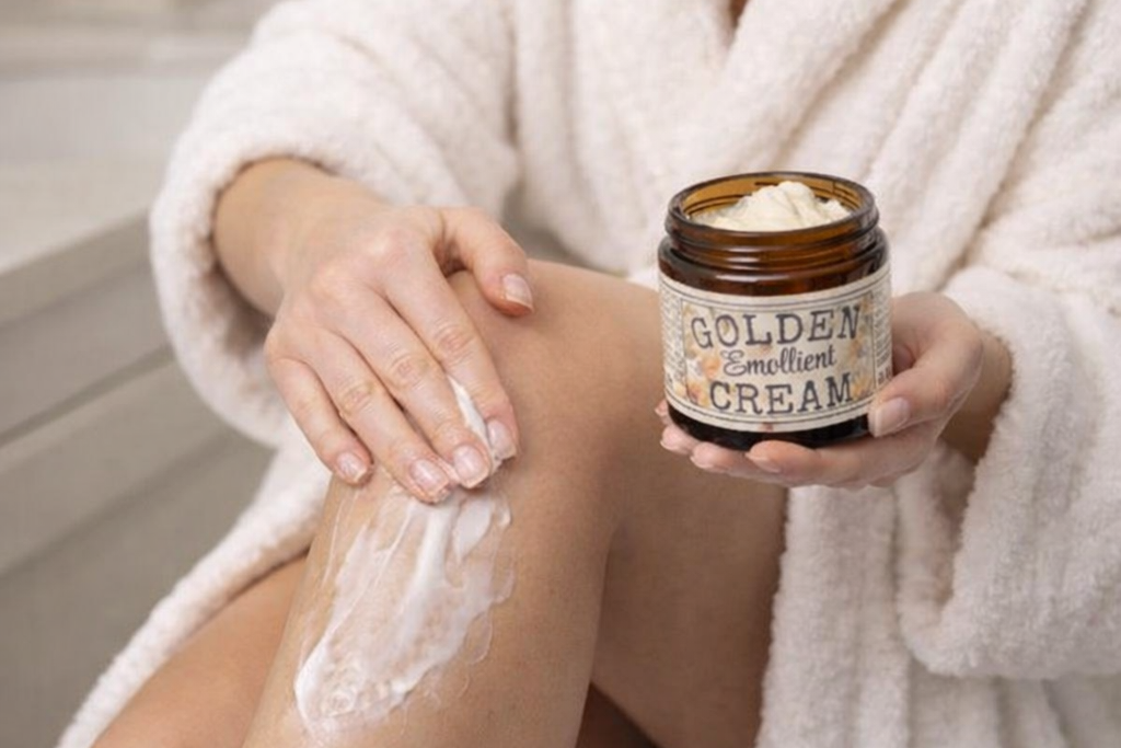 Woman in a bathrobe applying Golden Emollient moisturizing and skin conditioning Cream to her leg in a bathroom setting.
